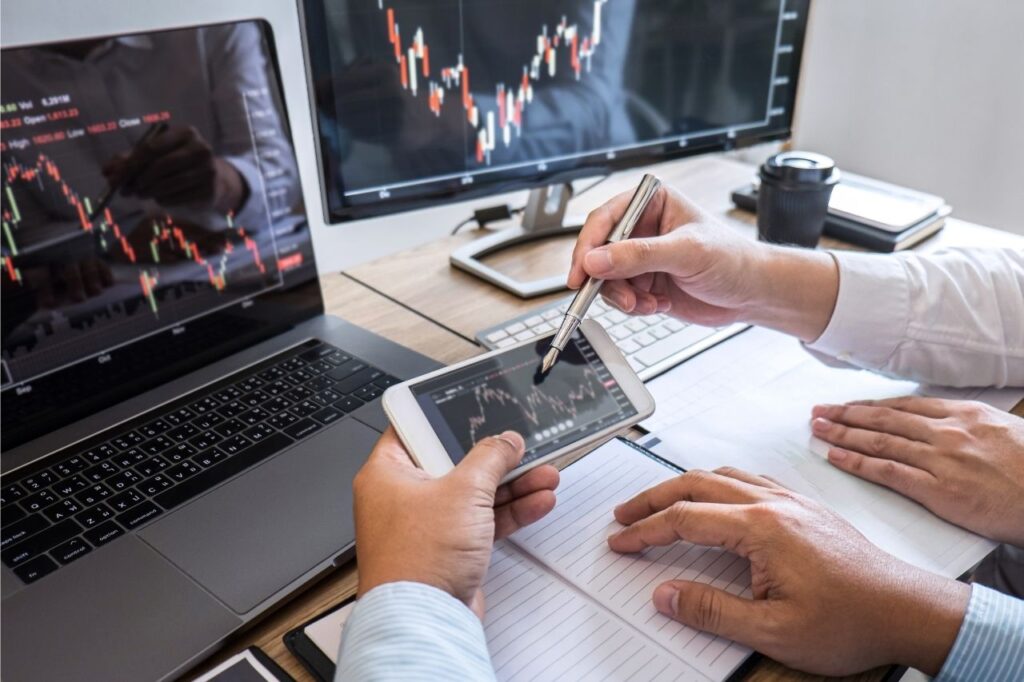 Na foto, dois homens analisando gráficos de investimentos no celular e no computador.